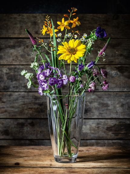 Die Kristallvase NACHTMANN Carre, gefüllt mit violetten, weißen und gelben Blumen auf einer hölzernen Anrichte.<br/>