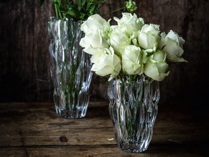 The small oval NACHTMANN Quartz vase filled with white roses in front of the tall NACHTMANN Quartz vase filled with white flowers on a sideboard made of dark wood.<br/>