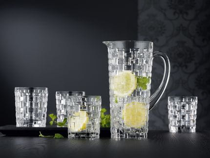 The NACHTMANN Bossa Nova jug and four tumblers on a black wooden table. The jug and one of the tumblers are filled with water, ice cubes, slices of lemon and mint sprigs.<br/>