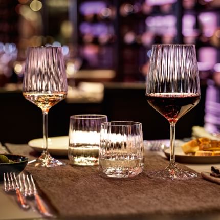 Die SPIEGELAU Lifestyle-Serie auf einem Restauranttisch in gemütlicher Abendatmosphäre. Violettes Licht schimmert durch die sanfte Wellenstruktur der gefüllten Weißwein- und Rotweingläser, Wassergläser und Karaffe.<br/>