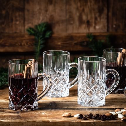 Das NACHTMANN Noblesse Teeglas mit Glühwein und Zimtstangen darin auf einer hölzernen Anrichte. Daneben stehen zwei leere und ein gefülltes Noblesse-Teeglas, Mandeln, Rosinen und Tannenzweige.<br/>