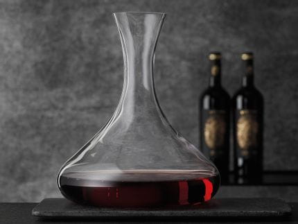 The NACHTMANN Vivendi premium decanter stands on a table, filled with red wine. Two black wine bottles can be seen in the background on the right.<br/>