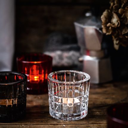 NACHTMANN Quadratische Teelichthalter in klarem Kristall, Rauch und Rot auf einem Holztisch. In ihnen funkeln Teelichter.<br/>