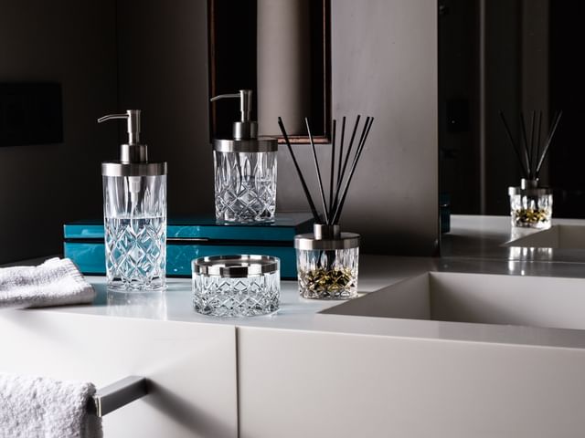 The NACHTMANN Spa Noblesse series on a bathroom sink in front of a mirror. The scent diffuser is filled with fragrance and black sticks. The small and the tall soap dispenser are filled with clear liquid. The storage jar is empty.<br/>