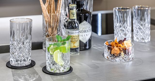 Il bicchiere longdrink NACHTMANN Noblesse Gold accanto al tumbler riempito con un cocktail a base di gin e tonic. Sullo sfondo un altro bicchiere longdrink riempito di crissini e una bottiglia di acqua tonica.<br/>