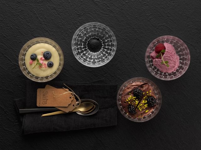 Four NACHTMANN Bossa Nova dip bowls, one filled with custard and blueberries on top, one with ice cream and a raspberry on top, one with chocolate cream with blackberries on top and one empty. To the right of it are spoons. All of them are on a black plate<br/>