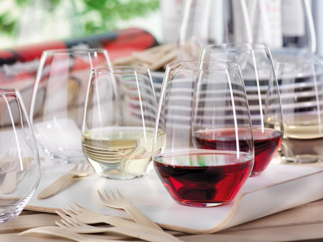 SPIEGELAU Authentis Casual tumblers on a serving tray on an outdoor table. Two of the stemless tumblers are filled with red wine, one with white wine. In the foreground are wooden forks and in the background a stack of plates and more tumblers.<br/>