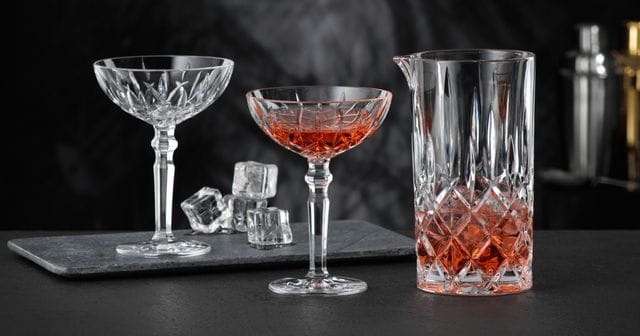 The NACHTMANN Noblesse pitcher filled with a red drink and ice cubes and two NACHTMANN Noblesse cocktail glasses, one of them filled with the same red drink. In the background there is the empty cocktail glass and ice cubes.<br/>