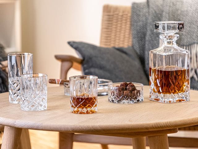 The NACHTMANN Noblesse tumbler with golden rim filled with whisky next to the bowl filled with snacks and the whisky decanter filled with whisky. All of them on a wooden table in front of a bench.<br/>