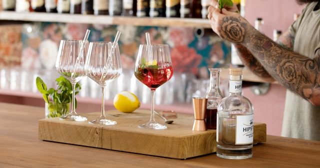 NACHTMANN Gin and Tonic glasses with glass straws on a bar counter. One is filled with a red Gin Cocktail with mint and a lemon zest. Behind the glasses is a lemon and a glass with mint sprigs. On the right are the hands of the bartender and bar equipment.