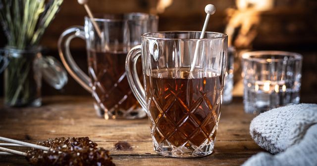 The NACHTMANN Noblesse tea mug with hot tea in it and rock candy. On the wooden table, there are more sticks with brown rock candy, a jar with lavender flowers, a second filled crystal tea mug and a shining tea light.<br/>