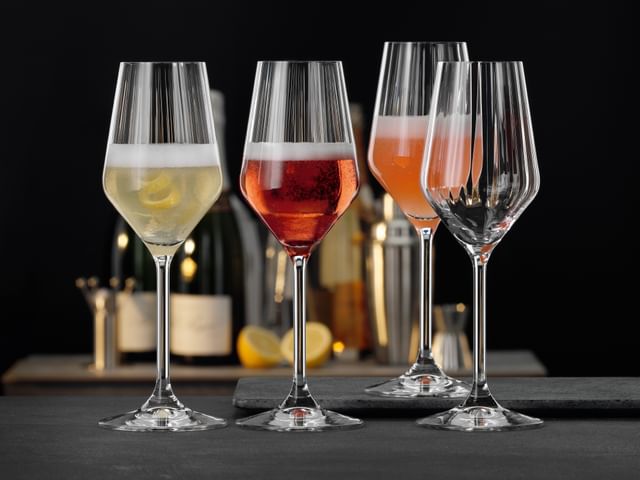 Four SPIEGELAU Lifestyle Champagne glasses. Three of them filled with a different Champagne cocktail, one of them empty. Behind them another table with Champagne bottles, lemons and bar equipment.<br/>