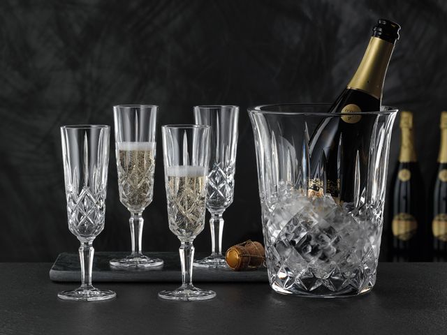 Four NACHTMANN Noblesse Champagne glasses two of which are filled with Champagne. Next to them is the NACHTMANN Noblesse Champagne cooler with ice cubes and an open bottle of Champagne inside.<br/>