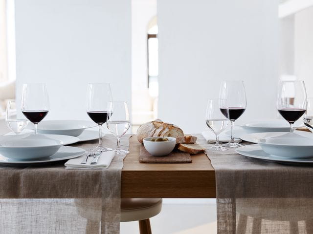 Das gefüllte SPIEGELAU Vino Grande Bordeaux Glas und Mineralwasserglas auf einem gedeckten Tisch. Auf den Platten befinden sich passende Suppenteller und daneben ist Besteck und Brot.<br/>