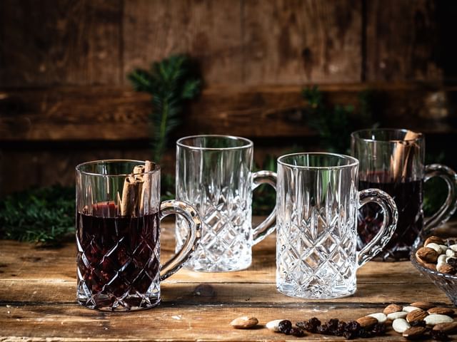 The NACHTMANN Noblesse tea glass with mulled wine and cinnamon sticks in it on a wooden sideboard. Next to it are two empty and one filled Noblesse tea glasses, almonds, raisins and pine branches.<br/>