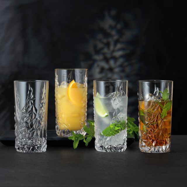 Vier NACHTMANN Sculpture Longdrink-Gläser auf einem Tisch, drei davon gefüllt mit verschiedenen alkoholischen und nicht-alkoholischen Getränken. Zwischen den Gläsern ist der Tisch mit Minzzweigen dekoriert.<br/>