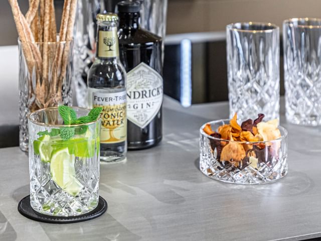 Il bicchiere longdrink NACHTMANN Noblesse Gold accanto al tumbler riempito con un cocktail a base di gin e tonic. Sullo sfondo un altro bicchiere longdrink riempito di crissini e una bottiglia di acqua tonica.<br/>