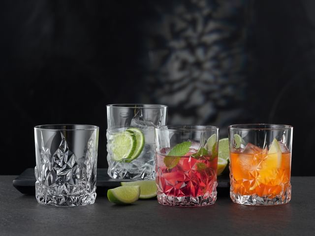 Four NACHTMANN Sculpture Whiskey tumblers on a table, three of them filled with different alcoholic and non-alcoholic drinks. Inbetween the glasses the table is decorated with slices of lime.<br/>